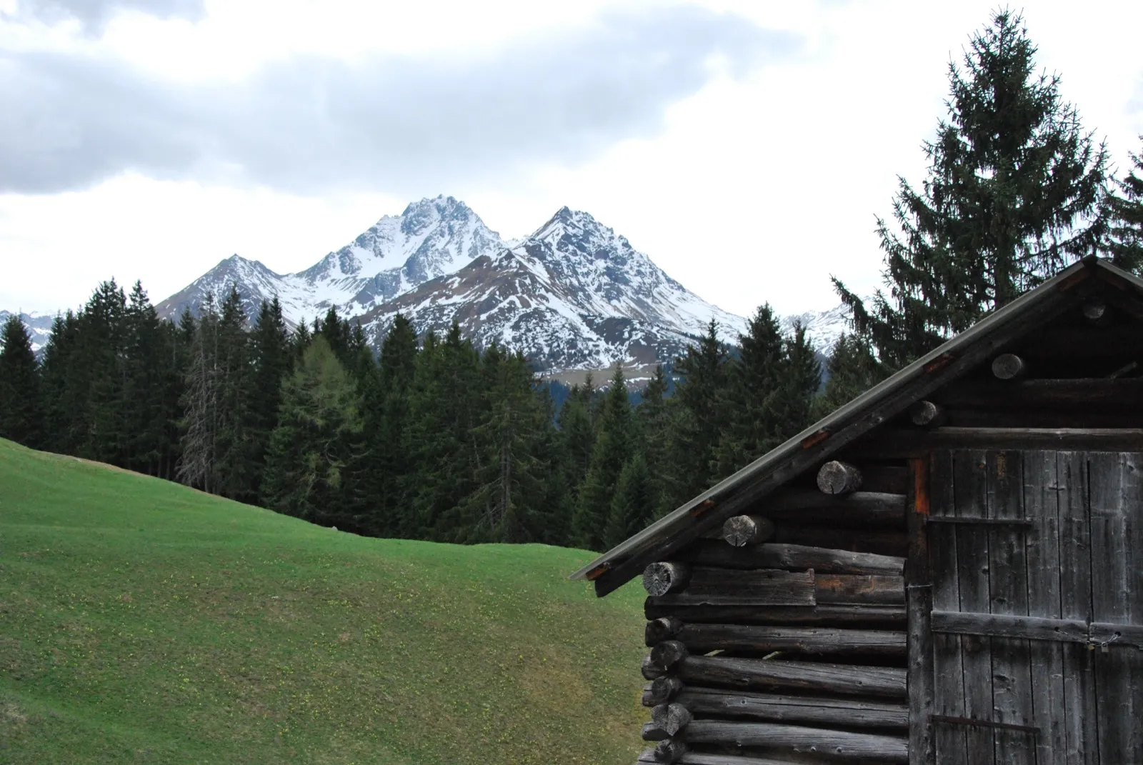 Wooden alpine hut on a green slope below snow covered mountains